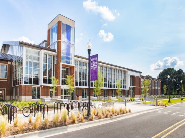 Modern university building with large glass windows, surrounded by trees and bike racks, under a blue sky.