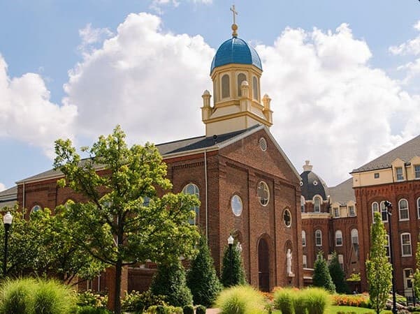 A brick building with a blue dome and cross on top, surrounded by greenery and trees, under a partly cloudy sky.