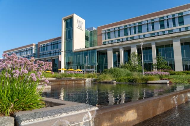 A modern college building with large glass windows, surrounded by greenery and flowers, reflecting in a water feature.